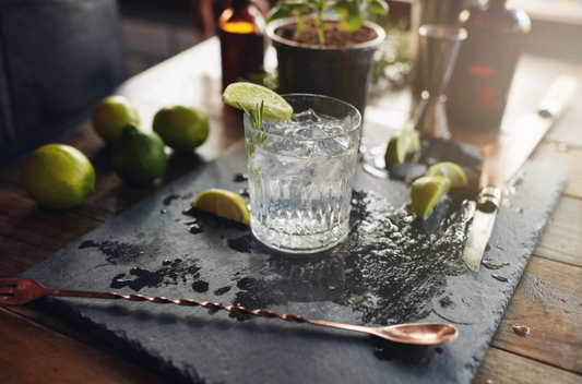 glass of gin over ice on bar with bartending tools and sliced limes