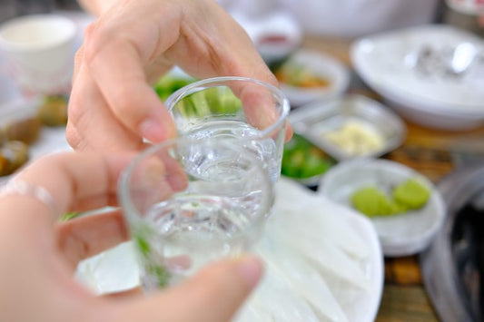 two hands clinking shot glasses of soju over dinner table