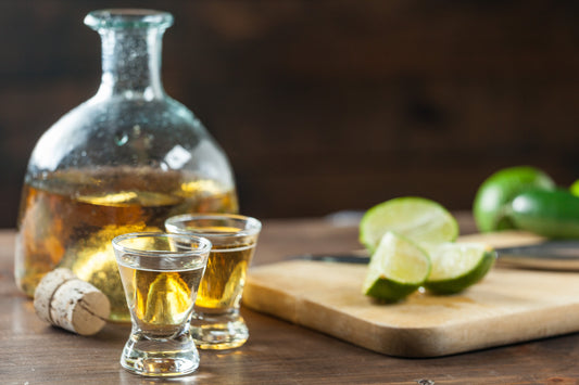 bottle of anejo tequila with two full shot glasses by sliced limes on wooden table with black background