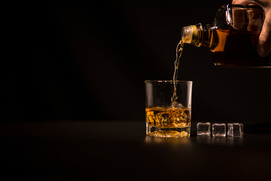 whiskey being poured from bottle into cocktai glass with ice cubes and black background