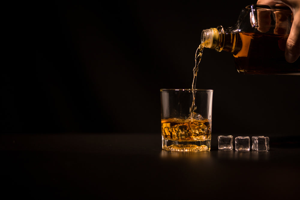 whiskey being poured from bottle into cocktai glass with ice cubes and black background