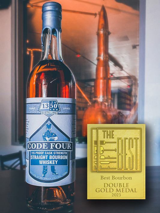 Double Gold Medal for The Fifty Best Bourbon 2023 sits to the left of a round bottle with gray label  in front of the "rocket still" in background. Hand-dipped silver wax drapes over the cork and neck of the bottle. An illustration of a police officer standing with arms crossed is behind the words “Code Four 115 Proof Cask Strength Straight Bourbon Whiskey”. The 1350 Distilling logo is centered at the top label.